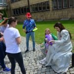 Młoda kobieta w sukni balowej siedzi na ławce z kilkuletnią dziewczynka. Przed nimi grupa młodzieży uśmiecha się do nich.