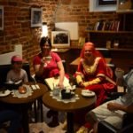 Mężczyzna, dwie kobiety oraz dwie małe dziewczynki siedzą przy kawiarnianych stolikach w pomieszczeniu ze ścianami ceglanymi o zamkowym wyglądzie.