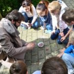 Siódemka dzieci wraz z dorosłym mężczyzna siedzą na kostce brukowej wokół naczynia z farbkami.