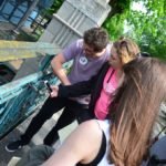 Dwie dziewczyny i trzech chłopaków stoją na moście, na którym przywieszone są kłódki zakochanych. Jedna z dziewczyn robi zdjęcie kłódce.