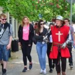 Grupa dziewczyn i chłopaków spaceruje ulica. Jeden z chłopaków ubrany jest w strój rycerski zakonu maltańskiego z czerwonym płaszczem i białym krzyżem.