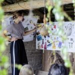 Kobieta stojąca przy arkuszach papieru przywieszonych na ścianie, na których są przyklejone zdjęcia, pocztówki i wycinki z gazet.